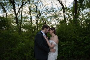 Bride and groom share romantic forehead touch surrounded by lush green trees at dusk.