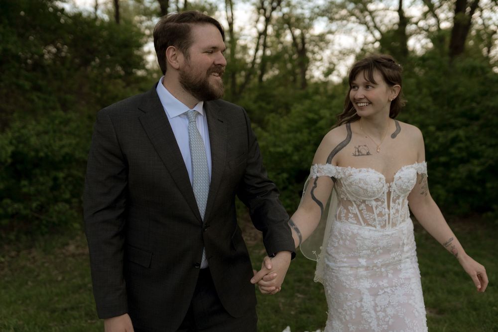 Newlywed couple holding hands and smiling in a garden, bride in lace gown and groom in dark suit.