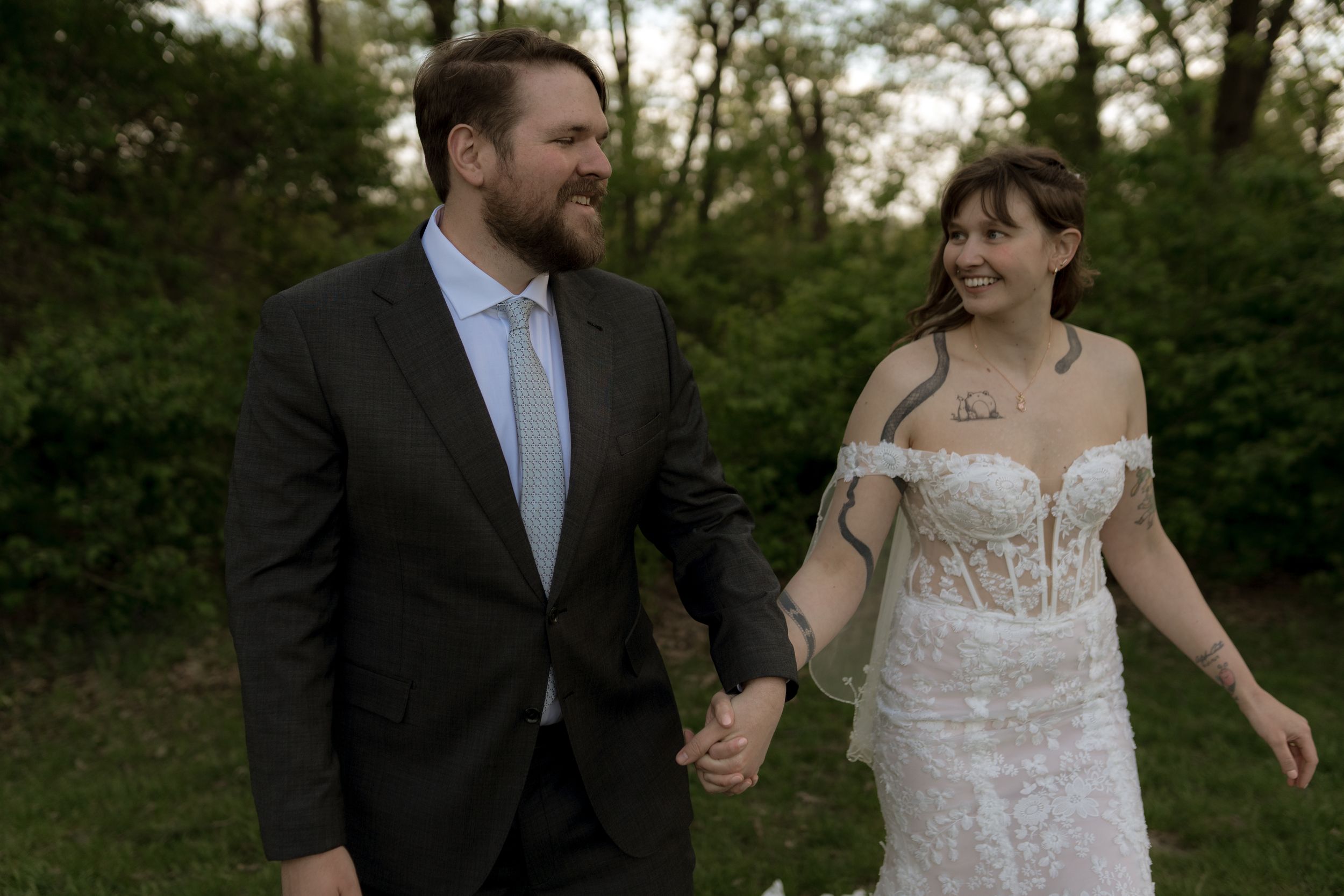 Newlywed couple holding hands and smiling in a garden, bride in lace gown and groom in dark suit.