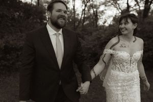 Bride in strapless lace gown and groom in suit hold hands smiling in black and white wedding portrait.