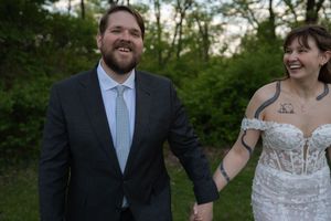 Smiling bride in strapless lace gown holds hands with groom in dark suit outdoors after wedding.