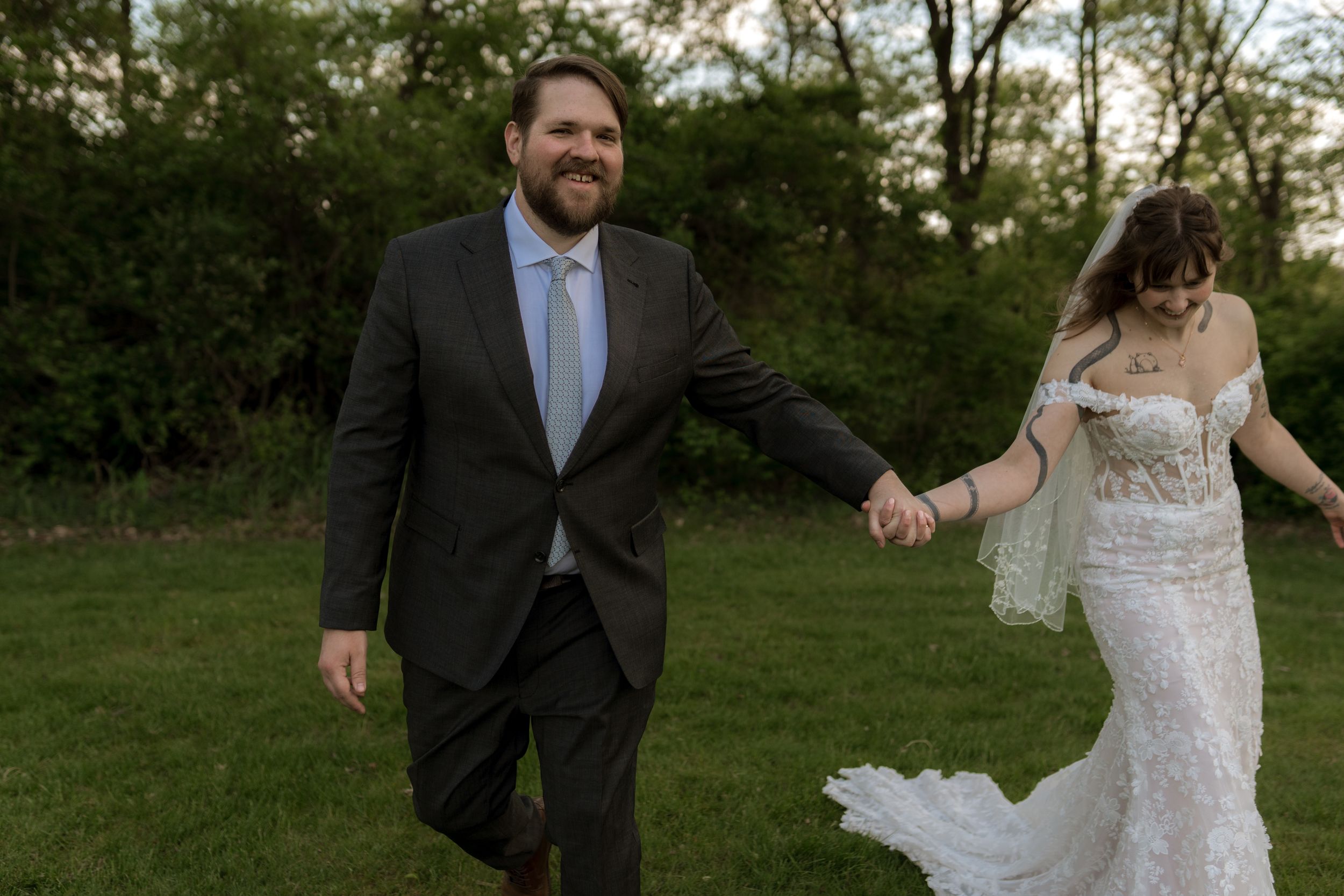 Groom in dark suit holds bride's hand walking across green lawn during wedding celebration.