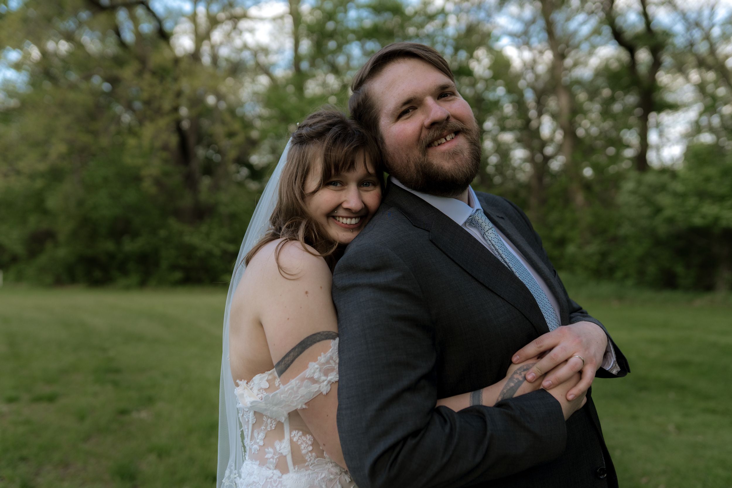 Happy newlyweds embrace outdoors, bride in lace backless gown with veil, groom in dark suit, surrounded by green trees.