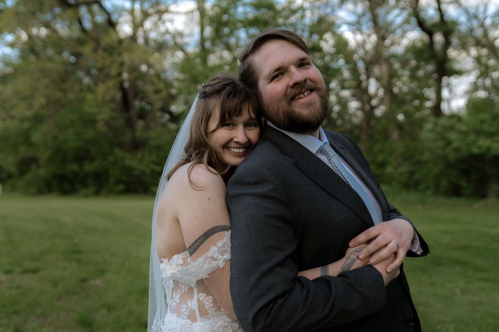 Happy newlyweds embrace outdoors, bride in lace backless gown with veil, groom in dark suit, surrounded by green trees.