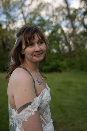 A smiling bride in an off-shoulder white lace wedding dress poses outdoors with soft bokeh greenery background.