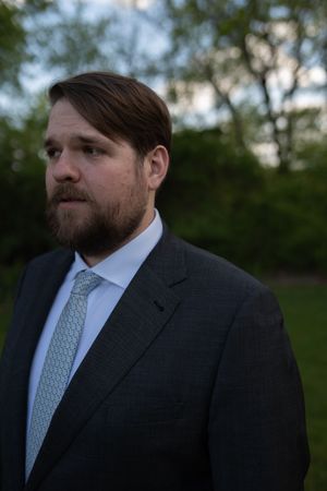 Bearded man in dark suit and teal tie standing outdoors with trees in background at dusk.