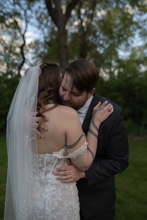 A bride in white lace gown and veil shares a tender embrace with groom in black suit in outdoor garden setting.