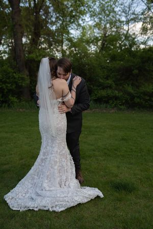 Groom embraces bride in elegant lace mermaid gown with long veil on green lawn.