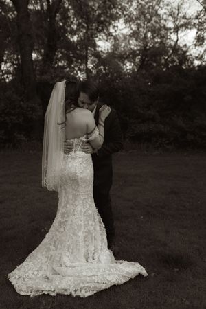 Black and white portrait of bride in lace gown with long veil embracing groom outdoors.
