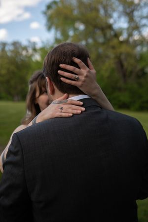 Close-up of bride's hands with rings resting on groom's shoulders and head during wedding.