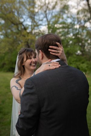 A joyful bride in white lace wedding dress and veil laughs while embracing her groom outdoors among green trees.