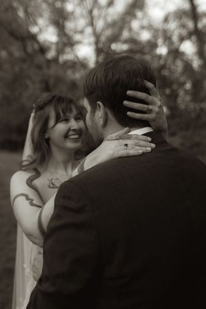 Black and white wedding photo of smiling bride in veil embracing groom outdoors among trees.