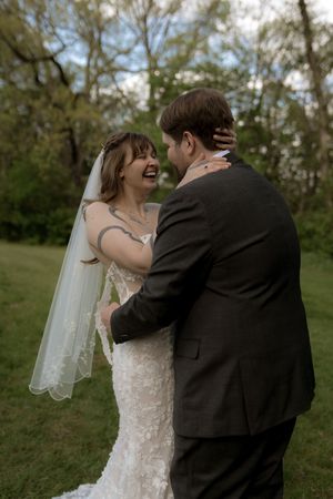 Joyful bride in lace wedding gown laughing while embracing groom in dark suit outdoors.