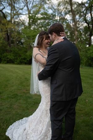 Bride and groom share an intimate embrace on green lawn, bride wearing lace mermaid gown with veil.