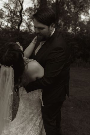 Romantic black and white photo of groom tenderly holding bride in lace dress in wooded setting.