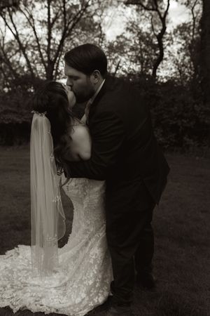 Bride and groom sharing a romantic dip kiss in black and white photo among trees at their wedding.