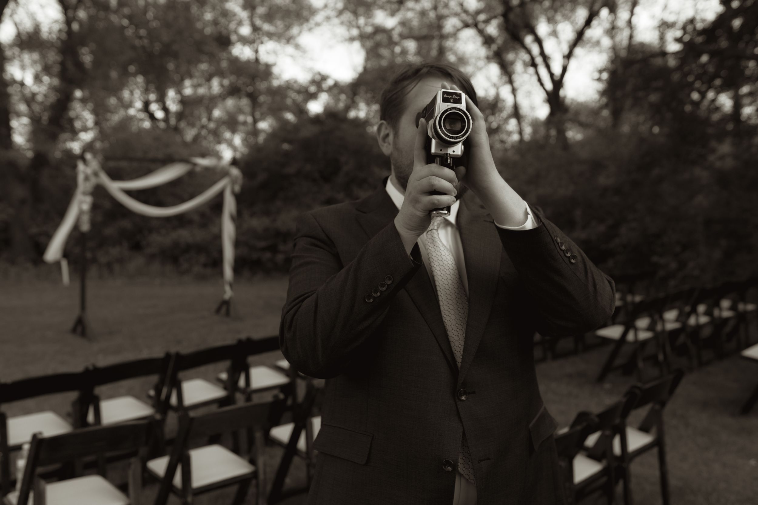Groom pointing vintage camera at photographer during outdoor wedding venue setup in black and white.