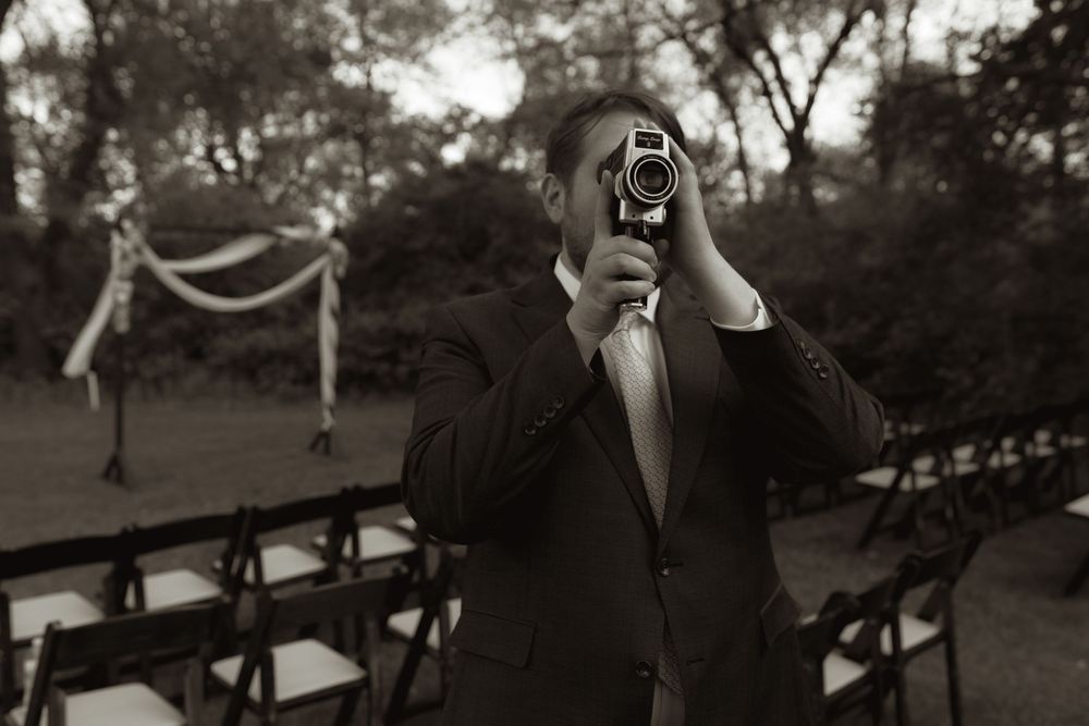 Groom pointing vintage camera at photographer during outdoor wedding venue setup in black and white.
