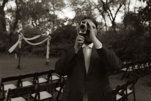 Groom in suit filming wedding ceremony with vintage camera in black and white outdoor setting.