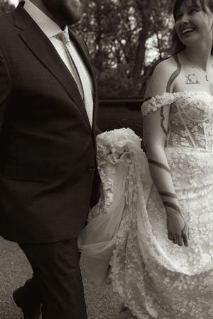 Black and white photo of bride in lace gown and groom in dark suit walking together at outdoor wedding.
