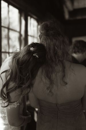 Two women in formal dresses share a tender moment, leaning heads together near a window in a black and white wedding photo.