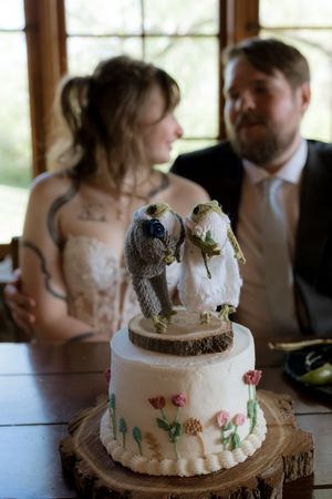 Wedding cake with bird topper on wood slab with couple blurred in background at rustic indoor reception.