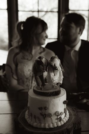 Black and white photo of bird cake toppers on wedding cake with couple in background.