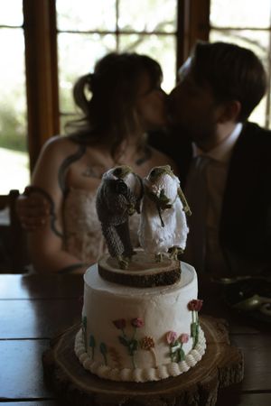 Wedding cake with bird toppers and floral decorations as couple kisses in background.