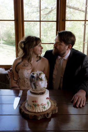 Bride and groom gaze at each other beside two-tier wedding cake with couple topper on rustic wood slice.