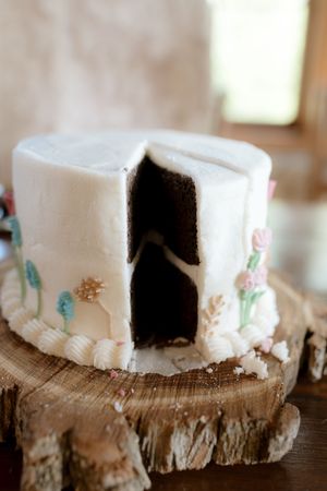 White frosted wedding cake with floral details on rustic wood stand, with a slice removed.