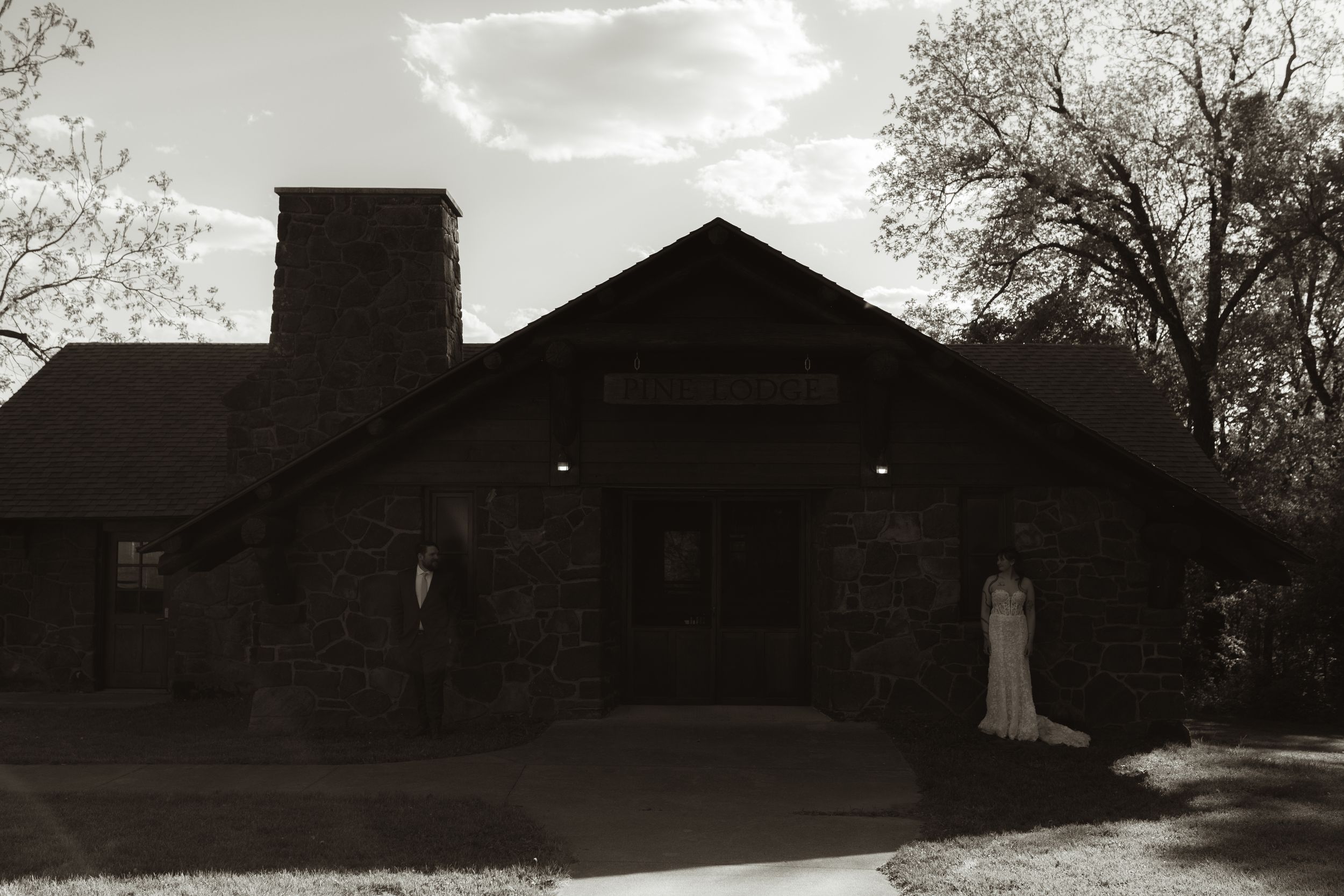 Black and white photo of a bride standing outside a rustic stone wedding venue with a chimney and trees.