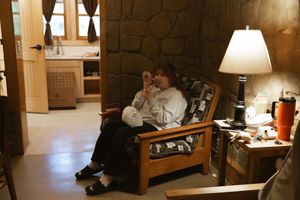 Person relaxing in a wooden armchair inside a rustic stone-walled cabin room with warm lamp lighting.