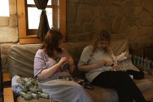 Two women sitting on a couch in a stone-walled cabin, working on crafts or knitting together.