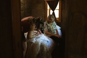 Bride in white lace wedding dress being assisted by attendants in a dimly lit rustic stone room.