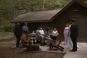 Wedding guests socializing outdoors near a rustic wooden cabin with a fire pit in a wooded setting.