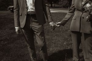 Couple holds hands walking through green grass in elegant suits, captured in romantic black and white wedding photography.