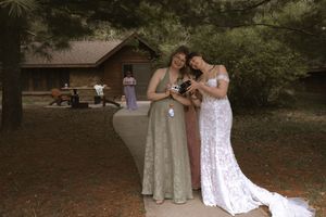 Two women in formal gowns, one white lace and one sage green, share a joyful moment outdoors at a woodland wedding venue.