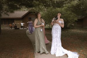 Bride in lace wedding gown and bridesmaid in sage green dress standing outdoors near rustic cabin.