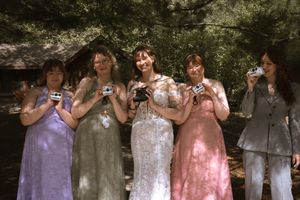 Bride and four bridesmaids smiling and holding cameras outdoors in colorful dresses.