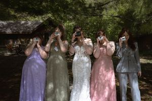 Bride and bridesmaids hiding faces behind cameras in fun outdoor wedding party photo.