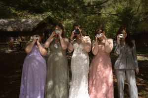 Bride in lace gown and bridesmaids playfully covering faces with cameras in sunny forest.