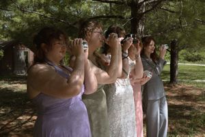 Bridesmaids in pastel gowns line up outdoors under pine trees taking photos at a wedding celebration.
