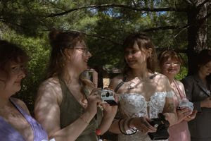 Bridesmaids laughing and taking photos outdoors under pine trees at a summer wedding celebration.