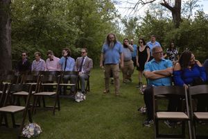 Wedding guests standing and seated on wooden chairs in an outdoor ceremony space surrounded by trees.