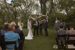 Outdoor wedding ceremony on green lawn with bride and groom under floral arch surrounded by wedding party and seated guests.