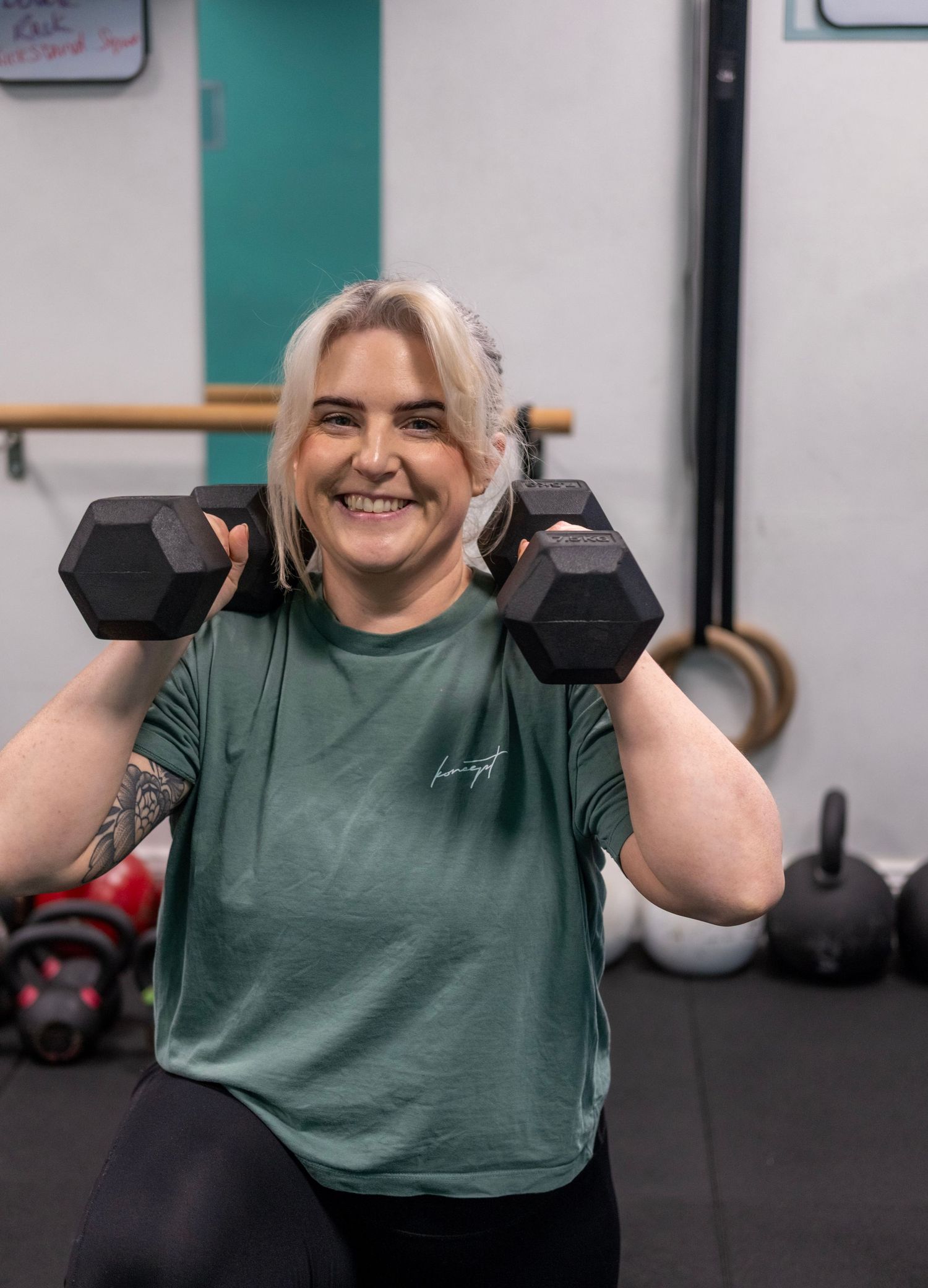 Smiling woman in green shirt lifting two black hex dumbbells at shoulders in a gym setting.
