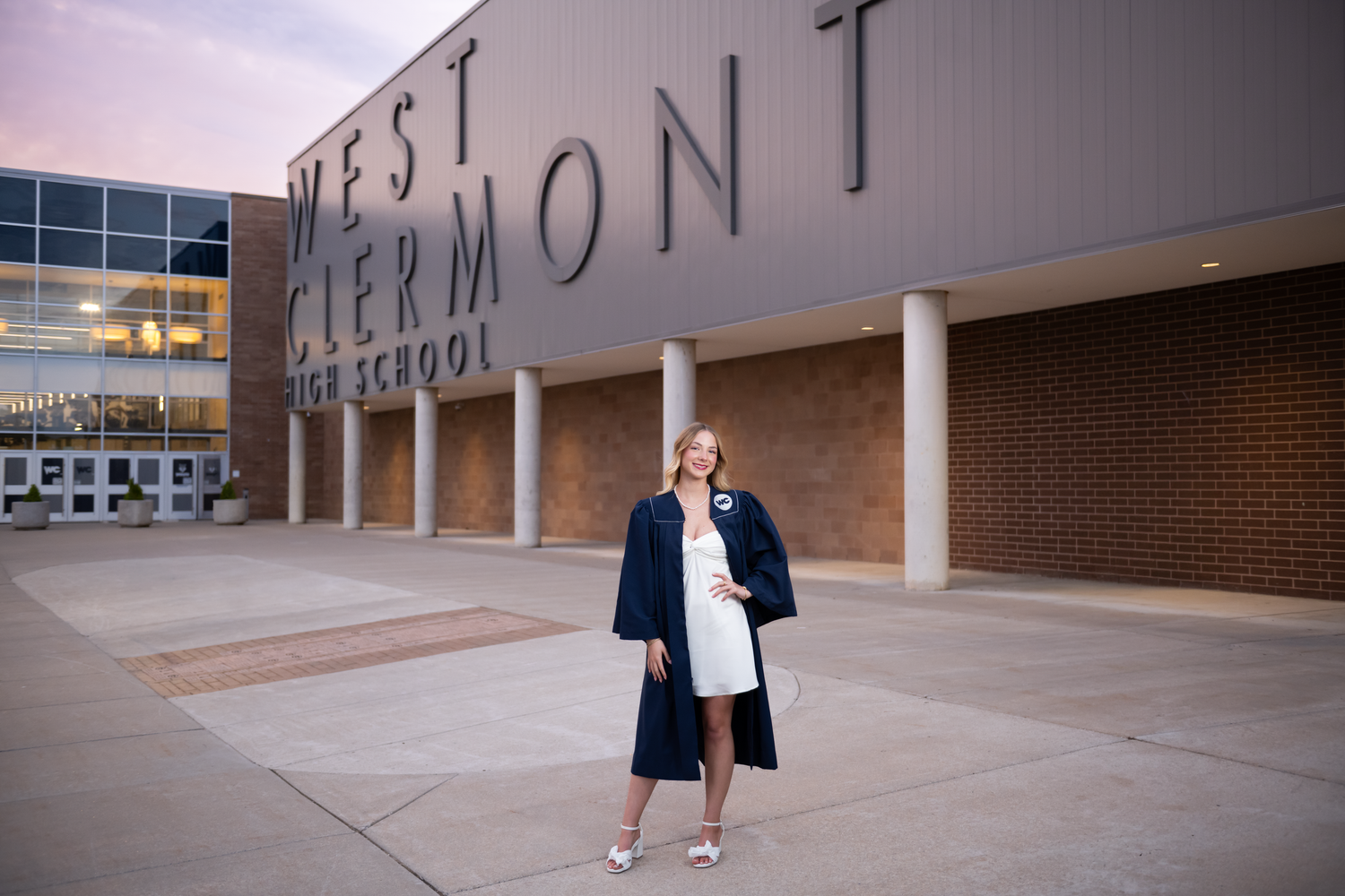 Graduate in cap and gown poses proudly outside West Clermont High School at dusk.