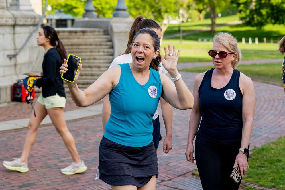 Three women walking outdoors on a brick path, one smiling and waving, wearing athletic wear on a sunny day.