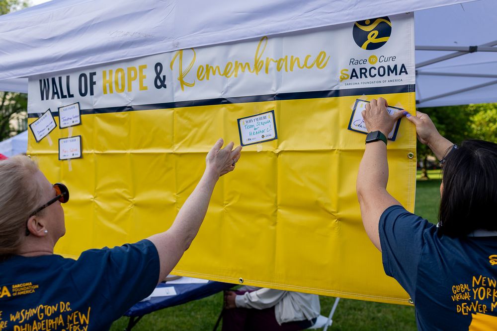 Volunteers post messages on a yellow Wall of Hope & Remembrance at a Sarcoma awareness event outdoors.