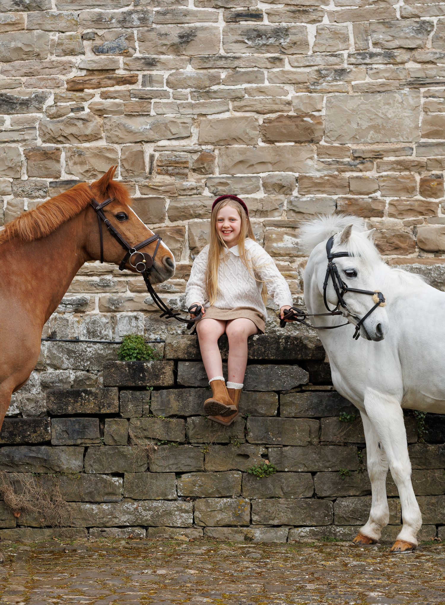 Young girl sitting on a stone wall between a chestnut horse and white pony, smiling outdoors.